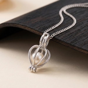 May include: A silver-colored pendant necklace featuring a pearl encased in a cage-like design. The pendant hangs from a delicate chain, resting on a dark wooden surface. The pearl is white and round, and the cage has a heart-shaped design.