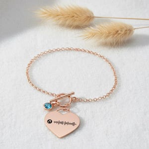 May include: Rose gold bracelet with a heart-shaped charm. The charm features a music note and a sound wave design. A small blue gemstone is set near the clasp. The bracelet is on a white surface with dried plants in the background.