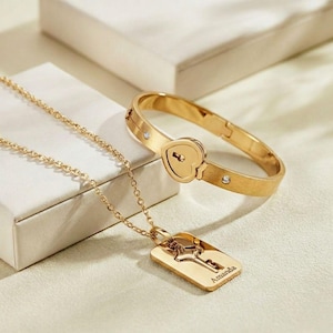 May include: A gold-coloured jewellery set comprising a necklace and a bracelet. The necklace features a rectangular pendant engraved with a key and the name "Amanda". The bracelet has a heart-shaped lock design and small, clear stones.