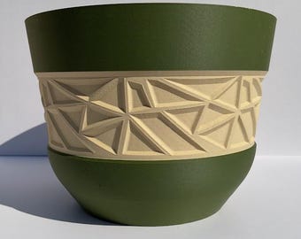 May include: A green and beige ceramic planter with a geometric design. The top and bottom sections are a solid green, while the middle band features a raised, angular pattern in beige. The planter has a rounded base and a wide opening.