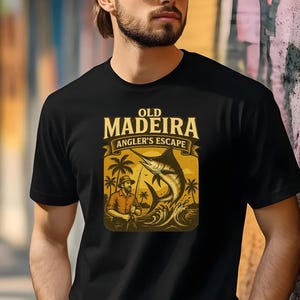 May include: Black t-shirt featuring a vintage-style graphic. The design includes the text "OLD MADEIRA ANGLER'S ESCAPE" above an illustration of a fisherman and a marlin jumping out of the water. Palm trees and waves complete the scene.