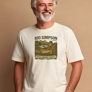 Może przedstawiać: Kremowa koszulka z grafiką pstrąga potokowego i wędkarza z napisem "RIO SIMPSON CHILE" i "WILD BROWN TROUT". Koszulka jest noszona przez osobę z siwymi włosami i brodą.