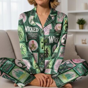 May include: Green pajama set with a repeating stamp design featuring the word "Wicked" and various illustrations. The set includes a button-down top and matching pants. The pajamas are worn by a person sitting on a couch.