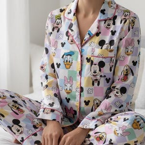 Op de afbeelding: Een Disney-pyjamaset met een kleurrijk ruitpatroon met Mickey Mouse, Donald Duck en Daisy Duck. De set bevat een overhemd met knopen en een borstzak en een bijpassende broek, afgewerkt met roze biezen.