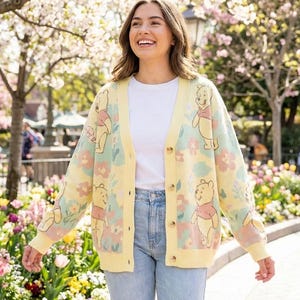 Op de afbeelding: Een gebreid vest met lange mouwen voorzien van een herhalend patroon van Winnie de Poeh-figuren, pastelroze bloemen en lichtblauw gebladerte op een lichtgele achtergrond, met een V-hals en een knoopsluiting aan de voorzijde met vier bruine knopen.