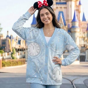 Puede incluir: Cárdigan azul claro con diseño de reloj y temática de Cenicienta. El cárdigan tiene botones en la parte delantera y mangas largas. El texto "Just before midnight" está impreso en el cárdigan. La persona de la imagen lleva una diadema con orejas de Minnie Mouse.