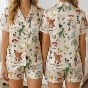 May include: Cream-colored pajama set featuring a print of Toy Story characters. The set includes a short-sleeved button-up shirt and matching shorts. The print includes characters like Woody, Buzz Lightyear, and Rex, along with the text "Bullseye", "Jesse", and "Buzz Lightyear".