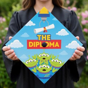 May include: A graduation cap with a blue sky background, white clouds, and green aliens. The cap features the text "THE DIPLOMA" in red and yellow, and a claw machine grabbing a diploma.