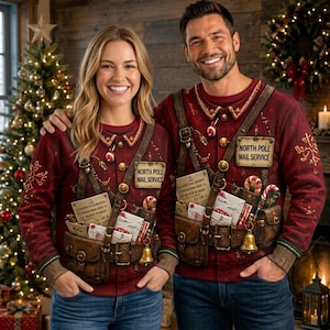 May include: Two matching red long-sleeve crewneck sweatshirts featuring a printed design of a brown leather mail carrier harness with pouches, a gold bell, candy canes, and a rectangular sign that reads North Pole Mail Service.