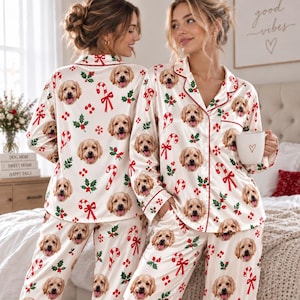 May include: A two-piece pajama set in a cream color featuring a repeating pattern of golden retriever dog faces, red candy canes, and green holly leaves. The long-sleeve button-down top has a notched collar with red piping and a matching pair of pants.