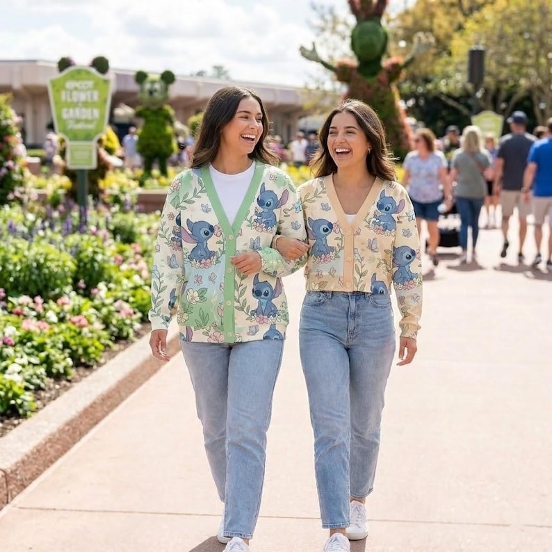 K&ouml;nnte beinhalten: Zwei Frauen tragen Stitch-Cardigans und hellblaue Jeans und gehen einen Weg entlang. Die Cardigans sind cremefarben mit einem Stitch-Charakterdruck und floralen Akzenten. Im Hintergrund ist ein Schild mit der Aufschrift "EPCOT Flower Garden" zu sehen.