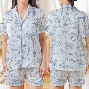 May include: White pajama set with a light blue outline pattern of seashells, palm trees, hearts, and other beach-themed illustrations. The text "Emily's Bachelorette Miami, FL 2025" is printed on the back of the shirt. The set includes a button-down shirt and shorts.