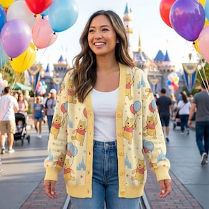 Może przedstawiać: Żółty kardigan z powtarzającym się wzorem Kubusia Puchatka trzymającego balony i sylwetki zamków. Kardigan ma zapinany na guziki przód i żółte wykończenia. Osoba nosi niebieskie dżinsy i biały top.