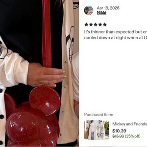 Op de afbeelding: Een wit vest met lange mouwen en een herhalende zwarte lijntekening van stripfiguurgezichten, gedragen over een zwart shirt, gecombineerd met een glanzende rode ronde schoudertas met afgeronde oren en een bijpassende rode riem.