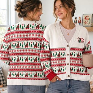 May include: A white knit cardigan featuring a repeating holiday pattern of red and green Christmas trees and castle silhouettes, with red and green horizontal stripes. The front includes a small embroidered graphic and the text EST. 1971.