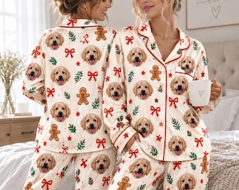 May include: A two-piece pajama set featuring a repeating pattern of golden retriever faces, gingerbread men, red bows, and green pine sprigs on a cream background. The long-sleeved button-down top has a notched collar and red piping trim.