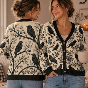 May include: A cream-colored V-neck cardigan featuring a repeating pattern of black crows perched on branches with blue berries. The garment has a black button-down front, black ribbed cuffs, and a black ribbed hem, worn over blue denim jeans.
