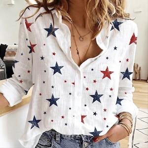 May include: A white long-sleeve button-down shirt featuring a scattered pattern of navy blue and red stars. The shirt has a classic collar, a front button placket, and long sleeves with cuffs, worn tucked into blue denim jeans.