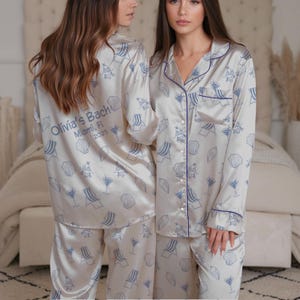 May include: Cream-coloured pyjama set with a beach-themed pattern in light blue. The set includes a button-down shirt and trousers, trimmed with navy blue piping. The back of the shirt has the text "Olivia's Bach Miami, Fl 2025".