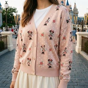 May include: A light pink cardigan sweater featuring a pattern of Minnie Mouse, red flowers, and white polka dots. The sweater has a V-neck, button closure, and ribbed cuffs and hem. The sleeves are long.