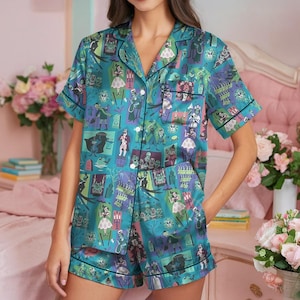 May include: Teal pajama set with a spooky, cartoon-style print featuring ghosts, tombstones, and other haunted house imagery. The short-sleeved top and shorts have black trim and a button-down front. The fabric has a silky sheen.