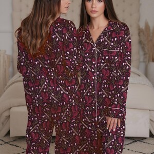 May include: Burgundy-colored pajama set with a floral pattern. The long-sleeved top and pants feature a white trim and a repeating design of stylized leaves and flowers. The pajamas are made of a soft, comfortable material.