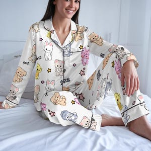 Peut inclure: Ensemble de pyjama blanc avec un motif r&eacute;p&eacute;t&eacute; d'animaux de dessins anim&eacute;s et d'&eacute;toiles. Le haut &agrave; manches longues et le pantalon ont une bordure noire et les lettres "SKZ". Le pyjama est en mati&egrave;re soyeuse.