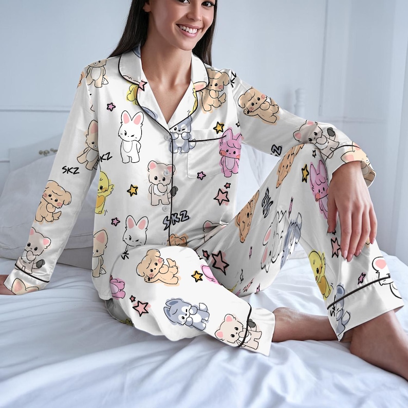 Peut inclure: Ensemble de pyjama blanc avec un motif r&eacute;p&eacute;t&eacute; d'animaux de dessins anim&eacute;s et d'&eacute;toiles. Le haut &agrave; manches longues et le pantalon ont une bordure noire et les lettres "SKZ". Le pyjama est en mati&egrave;re soyeuse.