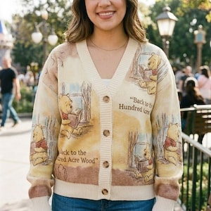 Op de afbeelding: Een beige vest met V-hals voorzien van een herhalend patroon van Winnie de Poeh die een boek leest. De gebreide stof toont de tekst Back to the Hundred Acre Wood in bruine letters. Het kledingstuk heeft een knoopsluiting met vijf bruine knopen.