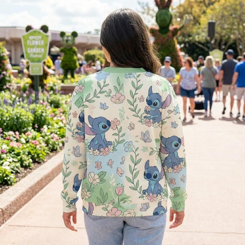 K&ouml;nnte beinhalten: Ein helles Sweatshirt mit einem sich wiederholenden Muster der Figur Stitch aus Lilo & Stitch, umgeben von Blumen und Schmetterlingen. Das Sweatshirt hat einen gr&uuml;nen Kragen und B&uuml;ndchen, und der Hintergrund ist eine Mischung aus hellgr&uuml;n und Creme.