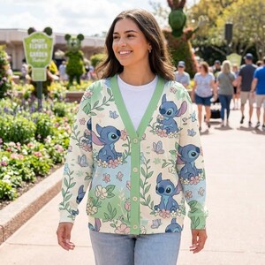 K&ouml;nnte beinhalten: Cremefarbene Strickjacke mit Stitch-Charakterdruck, umgeben von Blumen und Schmetterlingen. Die Strickjacke hat einen gr&uuml;nen Besatz und Kn&ouml;pfe. Der Hintergrund zeigt eine Gartenszene mit Menschen, die spazieren gehen.