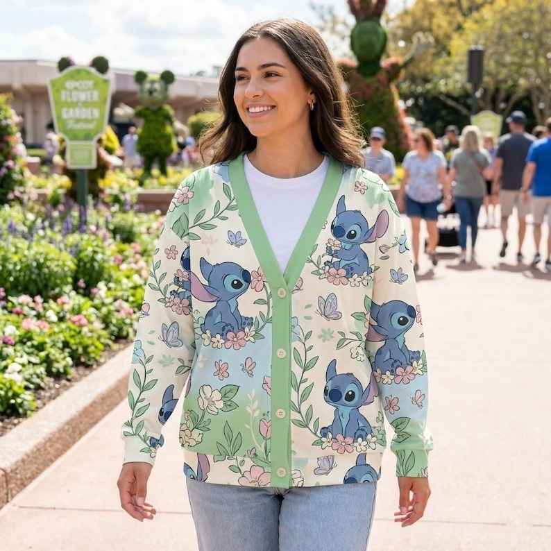 K&ouml;nnte beinhalten: Cremefarbene Strickjacke mit Stitch-Charakterdruck, umgeben von Blumen und Schmetterlingen. Die Strickjacke hat einen gr&uuml;nen Besatz und Kn&ouml;pfe. Der Hintergrund zeigt eine Gartenszene mit Menschen, die spazieren gehen.
