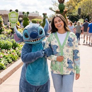 K&ouml;nnte beinhalten: Eine Frau tr&auml;gt einen cremefarbenen Cardigan mit Stitch-Muster, kombiniert mit einem wei&szlig;en Shirt und blauen Jeans. Der Cardigan zeigt Stitch-Illustrationen, Blumen und gr&uuml;ne Akzente. Eine Stitch-Figur steht neben der Frau.
