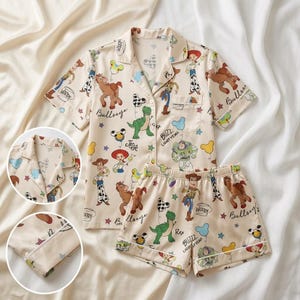 May include: Cream-colored pajama set featuring a button-down short-sleeved shirt and matching shorts. The set is adorned with cartoon character illustrations, including Woody, Buzz Lightyear, and other characters. The pajama set has white trim.