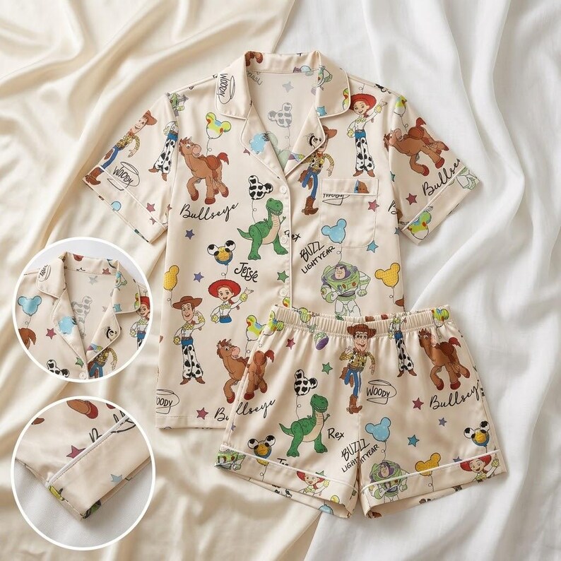 May include: Cream-colored pajama set featuring a button-down short-sleeved shirt and matching shorts. The set is adorned with cartoon character illustrations, including Woody, Buzz Lightyear, and other characters. The pajama set has white trim.
