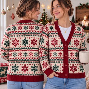 May include: A cream-colored V-neck cardigan featuring a festive red and green Nordic-inspired snowflake pattern. The garment has a solid red button-down placket, red cuffs, and a red hem. The knit fabric displays repeating geometric and snowflake motifs.