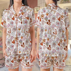 May include: A white pajama set with a repeating pattern of cartoon characters, cameras, coffee cups, and castle illustrations. The set includes a short-sleeved button-down shirt and matching shorts, both trimmed in a reddish-brown colour.