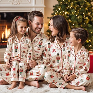 May include: Matching cream-colored pajama sets featuring a repeating pattern of nutcrackers, gift boxes, and rocking horses with red piping. The shirts have button-down fronts and collars, with the names Emma and Noah embroidered in red script.