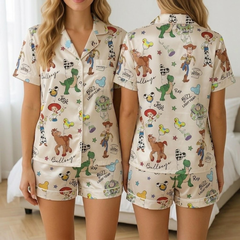 May include: Cream-colored pajama set featuring a print of Toy Story characters. The set includes a short-sleeved button-up shirt and matching shorts. The print includes characters like Woody, Buzz Lightyear, and Rex, along with the text "Bullseye", "Jesse", and "Buzz Lightyear".