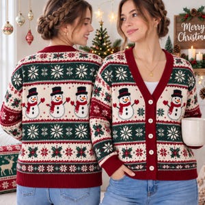 May include: A long-sleeve V-neck cardigan featuring a festive holiday pattern with snowmen wearing black top hats and red scarves, accented by green and white snowflake and holly motifs on a cream background with deep red trim and buttons.