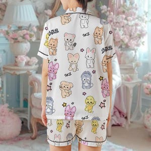 Peut inclure: Ensemble de pyjama blanc avec un motif r&eacute;p&eacute;t&eacute; d'animaux de dessins anim&eacute;s de diff&eacute;rentes couleurs, dont des ours, des lapins et des chiens. L'ensemble pr&eacute;sente une bordure noire et le texte "SKZ". Le haut &agrave; manches courtes et le short cr&eacute;ent un ensemble de v&ecirc;tements de nuit coordonn&eacute;s.
