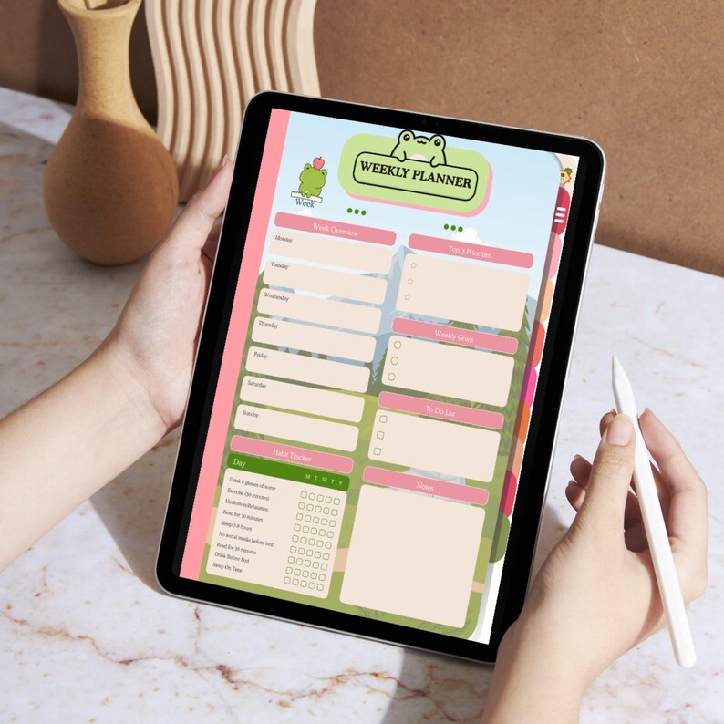 Op de afbeelding: Een digitale weekplanner op een tablet. De planner heeft een kikkerafbeelding en de tekst "WEEKLY PLANNER". De indeling omvat secties voor dagelijkse taken, notities en een gewoontetracker, met een roze en groen kleurenschema.