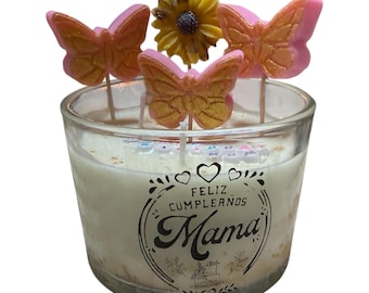 Puede incluir: Una vela de vidrio transparente con un interior de cera blanca. La vela está decorada con tres adornos de mariposas y una flor amarilla. La vela tiene las palabras "Feliz Cumpleaños Mama" impresas en el vidrio.