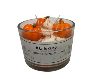 May include: A clear glass candle holder with a white label that reads "KC luxury Pumpkin Spice Luxe Scented Candles." The candle is filled with white wax and topped with three orange pumpkin decorations.
