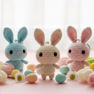 May include: Three handmade crochet bunny keychains in blue, beige, and pink. Each bunny has black eyes and pink accents. The scene includes pastel-colored eggs, flowers, and tulips, creating a spring or Easter theme.