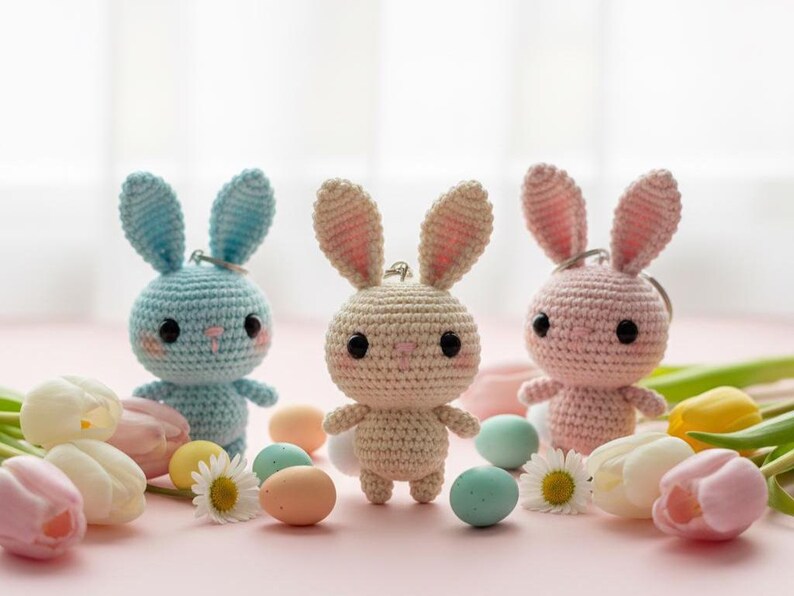 May include: Three handmade crochet bunny keychains in blue, beige, and pink. Each bunny has black eyes and pink accents. The scene includes pastel-colored eggs, flowers, and tulips, creating a spring or Easter theme.