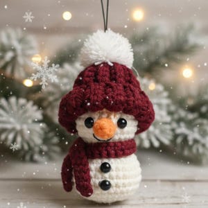 May include: A handmade crochet snowman ornament with a burgundy hat, scarf, and black button details. The snowman has a white body, an orange carrot nose, and a white pom-pom on top of the hat. The ornament is suspended by a black string.