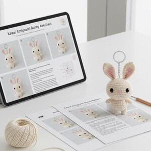 May include: A crocheted bunny keychain with pink-lined ears and black eyes sits on a white surface next to a crochet pattern and a ball of yarn. A tablet displays the pattern, titled "Kawaii Amigurumi Bunny Keechain."