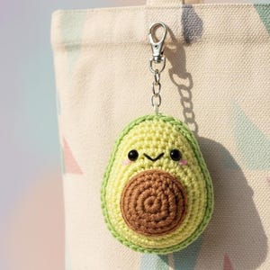 Può includere: Un portachiavi a forma di avocado all'uncinetto con un volto sorridente e un nocciolo marrone. L'avocado è verde e giallo, attaccato a un moschettone e una catena argentati. Il portachiavi è appeso a una borsa di tela.