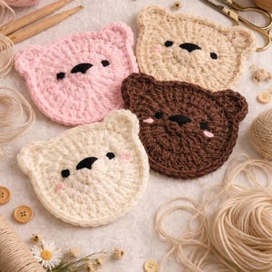 Puede incluir: Cuatro posavasos de oso de ganchillo hechos a mano en rosa, beige, marrón y blanco. Cada posavasos presenta una cara de oso con ojos y nariz negros y mejillas rosas. Hilo, botones y tijeras están esparcidos alrededor.